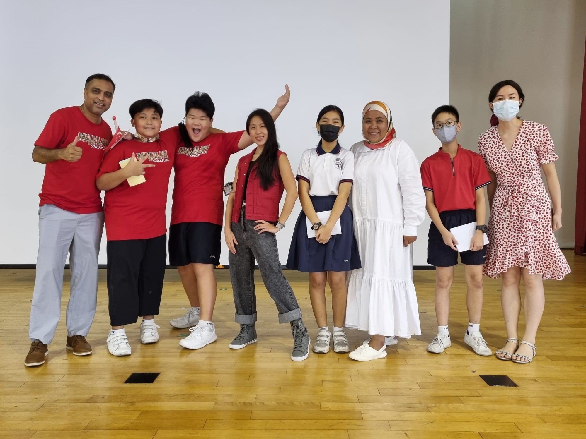 Eight people pose together on a wooden floor in front of a white screen, some wearing red shirts and face masks.